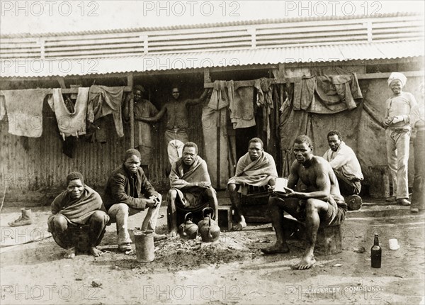 Off-duty diamond mine workers. African workers from the De Beers diamond mine rest outside their living quarters at the Kimberley compound. They sit around the remains of a fire and drink tea in front of a long, low tin-roofed shed. Kimberley, South Africa, circa 1896. South Africa, Central Africa, Oceania.