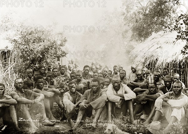 Acutt and Crewes labourers. A number of hired labourers sit on the ground in a scrubland clearing at Acutt and Crewe's labour depot near Bulawayo. They are barefoot and bareheaded, but most wear clothes of European origin. Matabeleland, Rhodesia (Matabeleland North, Zimbabwe), circa 1897., Matabeleland North, Zimbabwe, Southern Africa, Africa.