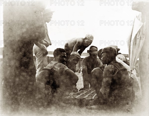 Off-duty diamond mine workers. African workers from the De Beers diamond mines sit around a small wooden frame, possibly a board game of some kind, at their living quarters in the Kimberley compound. Kimberley, South Africa, circa 1896. Kimberley, North Cape, South Africa, Southern Africa, Africa.