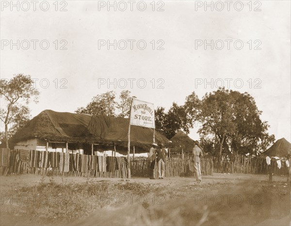 Mangwe Hotel and Store. Mangwe Hotel and Store, a single-storey thatched building surrounded by a wooden fence, over which are hung a number of striped towels or mats. Mangwe, Matabeleland, Rhodesia (Matabeleland South, Zimbabwe), circa 1896. Mangwe, Matabeleland South, Zimbabwe, Southern Africa, Africa.