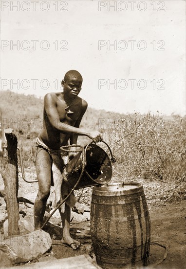 Collecting water at a village well. A Matabele (Ndebele) man pours water from a bucket into a barrel at a village well or water pump. Matabeleland, Rhodesia (Matabeleland North, Zimbabwe), circa 1897., Matabeleland North, Zimbabwe, Southern Africa, Africa.