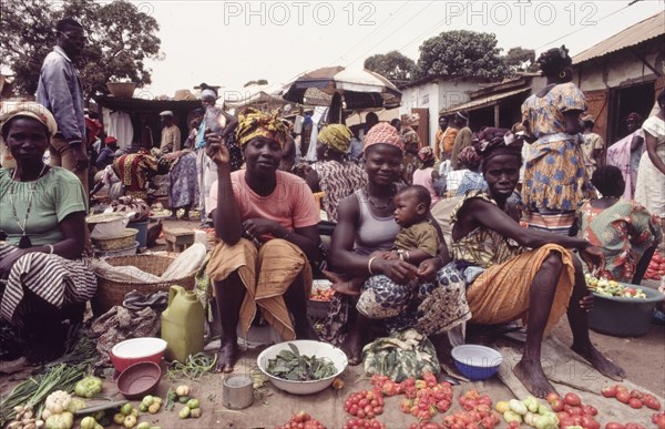 Female street traders at Banjul market. Female traders sell fruit and vegetables at a bustling outdoor market in Banjul. One of the women balances a baby on her knee, whilst another holds up a smoked fish for the camera. Banjul, Gambia, circa 1985. Banjul, Banjul City, Gambia, Western Africa, Africa.