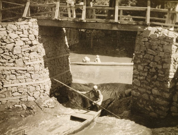 Shooting the rapids after the flood'. A man steers a canoe beneath a footbridge through rapids that have occurred as a result of a flood. Srinagar, Jammu and Kashmir State, India, circa 1927. Srinagar, Jammu and Kashmir, India, Southern Asia, Asia.