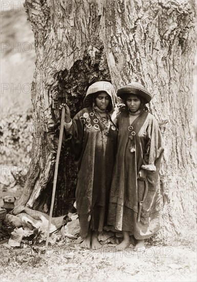 Two women from Kashmir. Portrait of two women from Kashmir, standing at the foot of a giant tree. Both women have their heads covered and wear long robes decorated with hoop brooches. One leans on a staff, whilst the other holds a sickle. Jammu and Kashmir State, India, circa 1925., Jammu and Kashmir, India, Southern Asia, Asia.