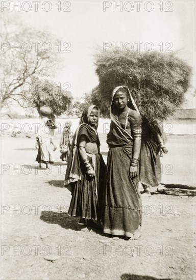 Two Indian women in Udaipur. Portrait of two young Indian women, dressed in traditional saris that cover their hair, and numerous bracelets and armbands. Behind them, several women struggle to carry large bundles of hay above their heads. Udaipur, Rajputana (Rajasthan), India, circa 1928. Udaipur, Rajasthan, India, Southern Asia, Asia.