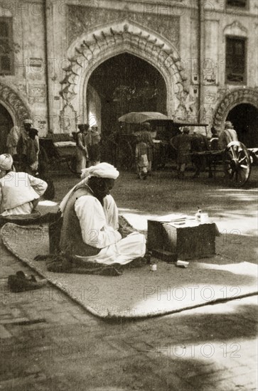 Letter-writing service. A street trader offering a letter-writing service sits on a rug on the ground, waiting for passing business at the side of a city street. North West Frontier Province, India (Pakistan), circa 1927., North West Frontier Province, Pakistan, Southern Asia, Asia.