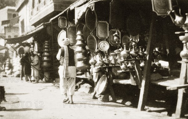 A copper bazaar. An Indian man in traditional dress walks past a row of market stalls crowded with goods at a copper bazaar. North West Frontier Province, India (Pakistan), circa 1927., North West Frontier Province, Pakistan, Southern Asia, Asia.