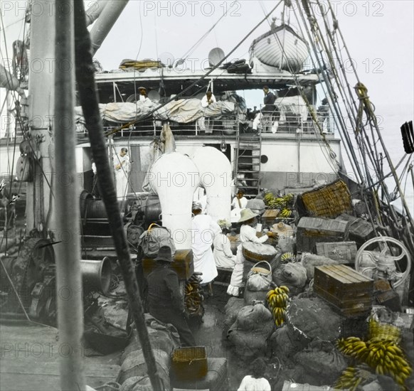 Bananas aboard the SS Chaleur. Jamaican traders travel aboard the SS Chaleur en route to Barbados. Hand-tinted detail draws attention to several bunches of bananas, a staple of Caribbean food, nestled amongst the cargo of sacks, baskets and trunks. Near Barbados, Caribbean, circa 1910., Caribbean, North America .