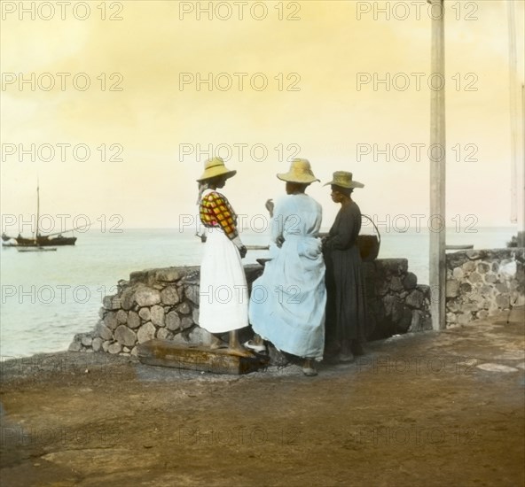 On the waterfront at Roseau. Three women in straw hats and dresses chat together on the waterfront at Roseau. Roseau, Dominica, circa 1910. Roseau, St Andrew (Dominica), Dominica, Caribbean, North America .