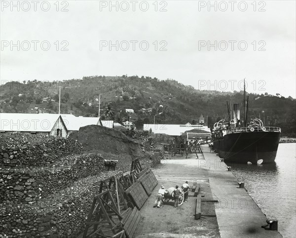 Unloading a cargo in St Lucia. A line of porters, carrying baskets on their heads, unload cargo from a ship moored at a harbour in St Lucia. They walk to and from the ship along a gangway, and appear to be carrying material, possibly coal, to a construction site. St Lucia, circa 1910. St Lucia, Caribbean, North America .