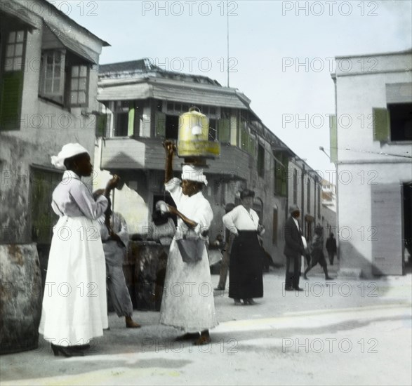 A soft drinks vendor in Barbados. A female street vendor balances a soft drinks decanter on her head and carries a metal pail over one arm as she approaches potential customers in the centre of town. Bridgetown, Barbados, circa 1910. Bridgetown, St Michael, Barbados, Caribbean, North America .