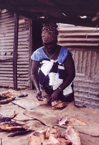Preparing fried fish at Bakau market. An African woman in traditional dress prepares dried fish on a stall at Bakau market. Bakau, Gambia, circa 1985. Bakau, West (Gambia), Gambia, Western Africa, Africa.