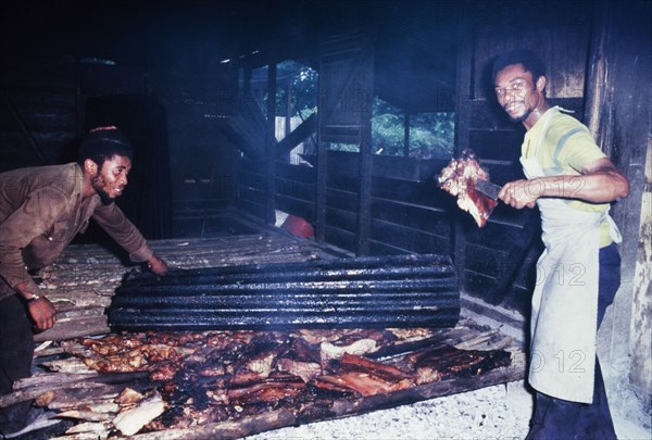 Preparing Jamaican pork jerk. An official Jamaican Tourist Board photograph features two men preparing pork jerk, a traditional Jamaican dish. The men turn over large pieces of pork, allowing it to cook evenly over a smoky fire. Port Antonio, Jamaica, circa 1985. Port Antonio, Portland, Jamaica, Caribbean, North America .