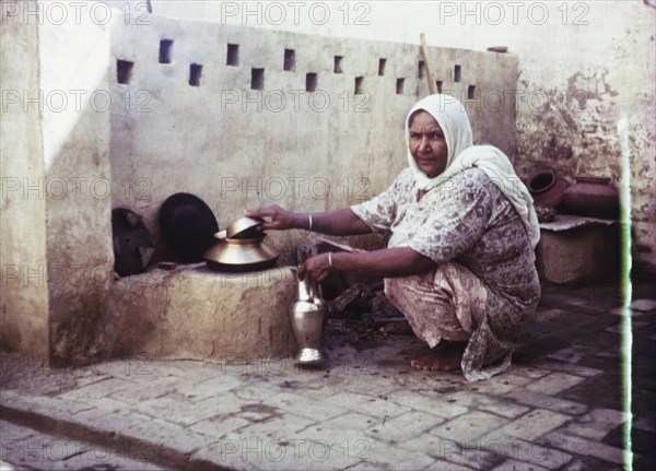 An Indian woman prepares to cook. A women squats beside a stone wall, preparing to cook with a metallic jug in one hand. India, circa 1985. India, Southern Asia, Asia.