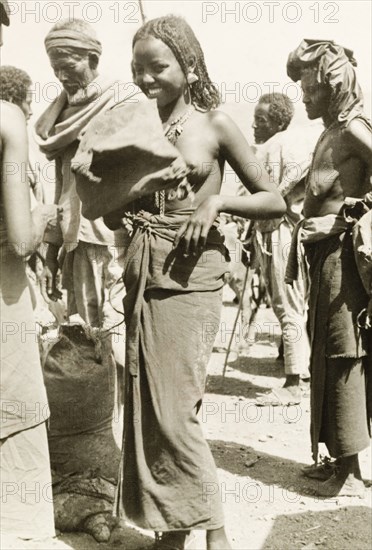 A young Eritrean woman. A young Eritrean woman is pictured during a moment of amusement at an outdoor camel market. Naked from the waist up, her hair is braided and she wears a decorative necklace and earrings. Eritrea, 1943. Eritrea, Eastern Africa, Africa.