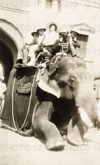 Europeans take an elephant ride, India. A group of smiling European people sit in a howdah on the back of an elephant as they prepare for a ride. The elephant's mahout (elephant handler) sits on its shoulders, 'ankusha' in hand. Lahore, Punjab, India (Pakistan), 3 April 1920. Lahore, Punjab, Pakistan, Southern Asia, Asia.