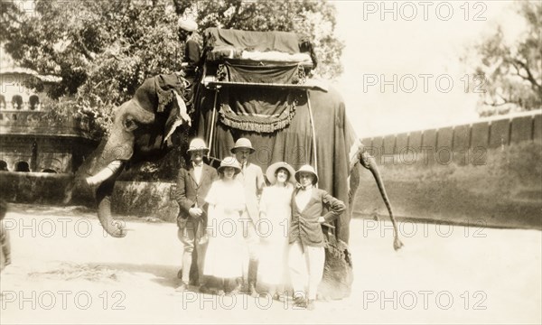 Europeans take an elephant ride. A group of European people pose for a photograph in front of an elephant saddled with a howdah, shortly before embarking on an elephant ride. The elephant's mahout (elephant handler) sits on its shoulders, 'ankusha' in hand. Lahore, Punjab, India (Pakistan), 3 April 1920. Lahore, Punjab, Pakistan, Southern Asia, Asia.