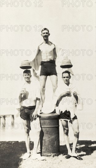 Hats off Brigadier Winthrop. Three European men pose for a comical photograph after a swim, wearing only swimming trunks with towels wrapped around their shoulders. A man identified as Brigadier Winthrop stands on a mooring post between his companions, holding their solatopi hats several inches above their heads. India, 1920. Kota, Rajasthan, India, Southern Asia, Asia.