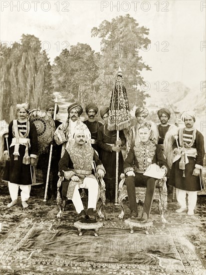 Sir Edward Maclagan and the Raja of Suket. Sir Edward Maclagan, Lieutenant Governor of the Punjab, and the Raja of Suket pose for a portrait against a painted backdrop. They sit on ornately decorated chairs, wearing full ceremonial dress, and are attended by Indian servants bearing parasols and horsehair fly whisks. Probably Suket, Rajputana (Rajasthan), India, 1920. Suket, Rajasthan, India, Southern Asia, Asia.