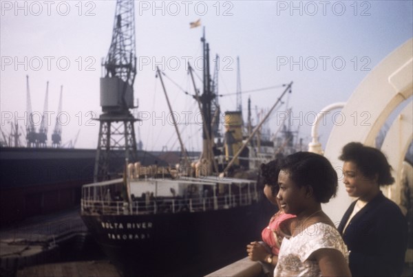 Shipping at Tilbury Docks. Three West African women gaze over the side of a ship moored at Tilbury Docks on the River Thames. A large steamship, 'River Volta', sits in harbour in the distance. Tilbury, England, September 1958. Tilbury, Essex, England (United Kingdom), Western Europe, Europe .