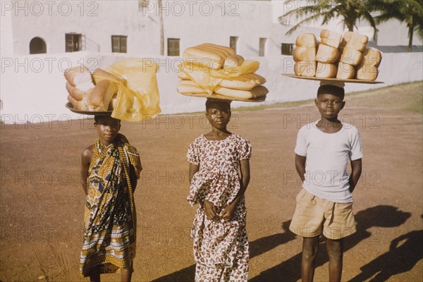 Breadsellers outside Elmina Castle. Three breadsellers pose for the camera, balancing large trays of bread on their heads outside the walls of Elmina Castle. Elmina, Ghana, circa 1959. Elmina, Central (Ghana), Ghana, Western Africa, Africa.