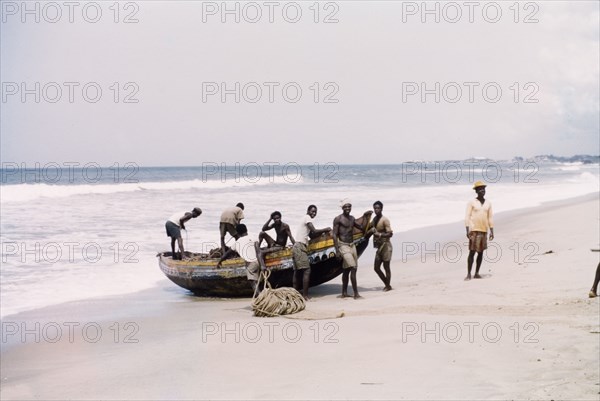 Fishing boat at Cape Coast. A group of men stand around a small fishing boat that is grounded on a beach. Near Cape Coast, Ghana, circa 1958. Cape Coast, Central (Ghana), Ghana, Western Africa, Africa.