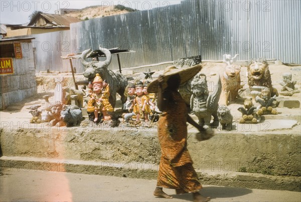 Ornaments at a roadside stall. A woman wearing a wide-brimmed sun hat passes by a roadside stall selling a variety of large stone ornaments. Cape Coast, Ghana, March 1958. Cape Coast, Central (Ghana), Ghana, Western Africa, Africa.