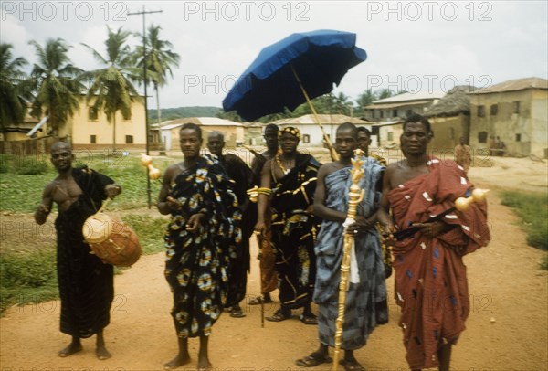 An Ghanaian chief and his retinue. A Ghanaian chief and his retinue process along a road in Anomabu. Members of the entourage wear traditional dress and carry various items including a golden staff, a ceremonial umbrella and a drum. Anomabu, Ghana, January 1958. Anomabu, Central (Ghana), Ghana, Western Africa, Africa.