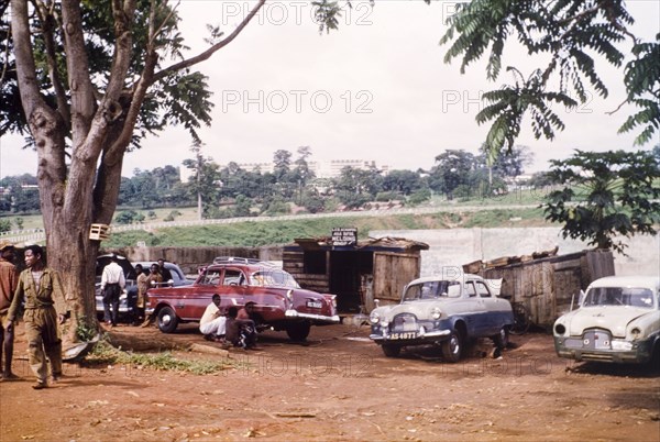 Car workshop in Kumasi. Mechanics work at a roadside car repair workshop. Kumasi Hospital is just visible in the distance. Kumasi, Ghana, circa May 1959. Kumasi, Ashanti, Ghana, Western Africa, Africa.