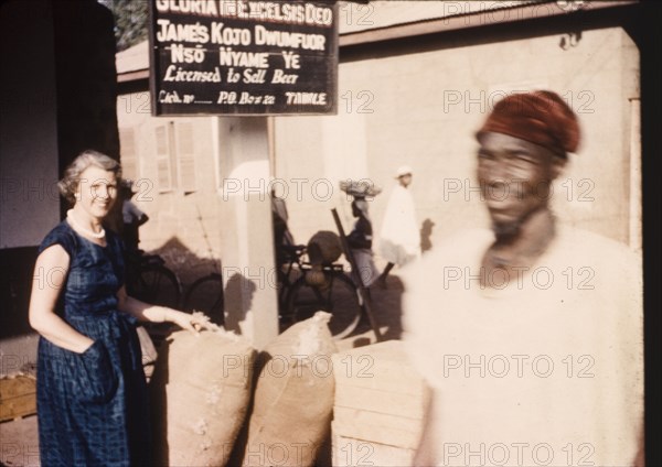 Street trader's stall in Tamale. A European woman inspects sacks at a street traders stall beneath a sign that reads: 'Gloria in Excelsis Deo, James Kojo Dwum Nso Nyame Ye, Licensed to Sell Beer'. Tamale, Ghana, circa 1958. Tamale, North (Ghana), Ghana, Western Africa, Africa.