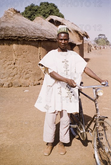 Ghanaian man with his bicycle. A Ghanaian man wearing traditional dress stands outside a mud-walled hut with his bicycle. Pong-Tamale, Ghana, circa 1961. Pong-Tamale, North (Ghana), Ghana, Western Africa, Africa.