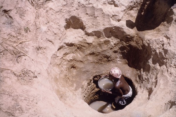 Waterhole near Tamale. A Ghanaian woman collects water in a pail from the bottom of a deeply-dug waterhole. Near Tamale, Ghana, circa 1961. Tamale, North (Ghana), Ghana, Western Africa, Africa.