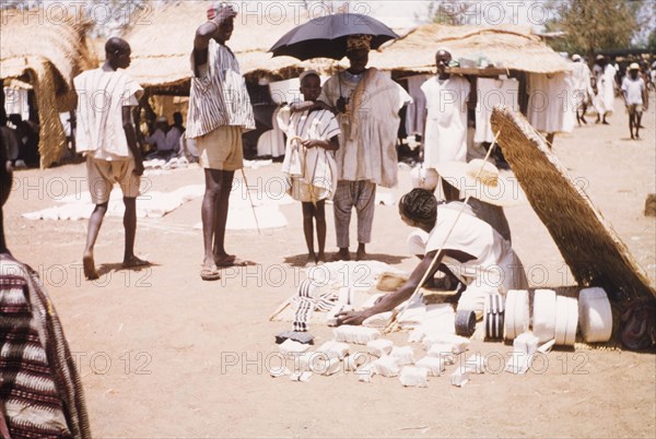 Woven cloth at Bawku market. A trader sells woven strip cloth from beneath a thatched sun shelter at Bawku market. Bawku, Ghana, circa March 1961. Bawku, Upper East, Ghana, Western Africa, Africa.