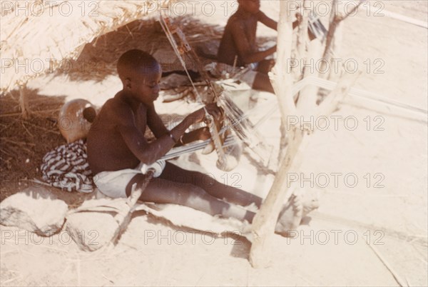 Boys weaving cloth at Pusiga, Ghana. Two young boys sit outdoors beneath thatched sun shelters as they weave strip cloth using heddle looms. Pusiga, Ghana, circa 1960. Pusiga, Upper East, Ghana, Western Africa, Africa.