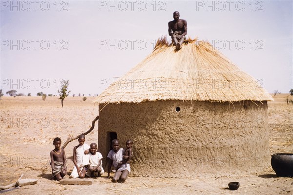 Roof thatcher in northern Ghana. A roof thatcher sits atop a round mud hut in Kuka. Thatchers in this arid part of far northern Ghana work with sheaves of straw, layering it thickly to provide a roof that is both waterproof and well insulated against the heat. Kuka, Ghana, circa March 1961. Kuka, Upper East, Ghana, Western Africa, Africa.