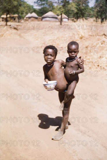 On the Gambaga-Walewale road'. A naked child carrying a bowl balances a toddler on his/her hip on the Gambaga-Walewale Road. Both children have distended bellies and may be suffering from malnutrition. Northern Ghana, circa 1960., North (Ghana), Ghana, Western Africa, Africa.