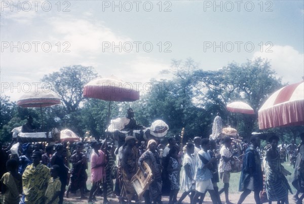 Procession at a Ngmayem festival. A procession of people passes through an open, grassy space at an annual Ngmayem harvest festival. Several Manya Krobo chiefs, accompanied by their 'okyeames' (linguists) who carry 'pomas' (linguists' staffs), are ferried along in palanquins shaded by umbrellas. Odumasi, Ghana, circa 1960. Odumasi, East (Ghana), Ghana, Western Africa, Africa.