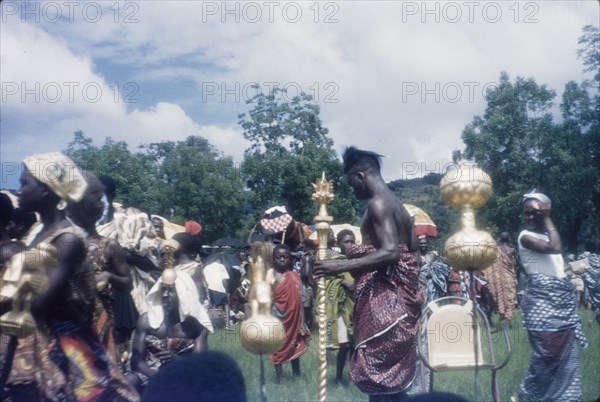The chief linguist of Mate Kole II. The chief 'okyeame' (linguist) of Mate Kole II (1910-1990), the Paramount Chief or Konor of Manya Krobo, carries a 'poma' (linguist's staff) through crowds celebrating at an annual Ngmayem harvest festival. This photograph captures him as he walks between the ornate handles of the Konor's ceremonial swords. Odumasi, Ghana, circa 1960. Odumasi, East (Ghana), Ghana, Western Africa, Africa.