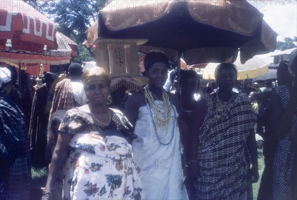 Notable women at a Ngmayem festival. Three finely dressed women, possibly the wives of Manya Krobo chiefs, stand beneath a ceremonial umbrella at an annual Ngmayem harvest festival. Behind them, a bearer carries a ceremonial stool above his head, a traditional Asante (Ashanti) symbol of a chief's leadership. Odumasi, Ghana, circa 1960. Odumasi, East (Ghana), Ghana, Western Africa, Africa.