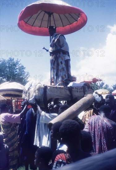 A Manya Krobo chief stands on a palanquin. A Manya Krobo chief stands up on a palanquin beneath a ceremonial umbrella as his retinue of bearers carries him through crowds at an annual Ngmayem harvest festival. Odumasi, Ghana, circa 1960. Odumasi, East (Ghana), Ghana, Western Africa, Africa.