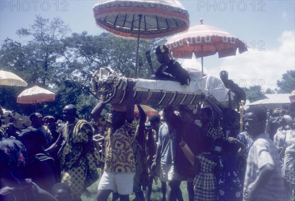 A Manya Krobo chief sits in a palanquin. A Manya Krobo chief sits in a palanquin beneath a ceremonial umbrella as his retinue of bearers carries him through crowds at an annual Ngmayem harvest festival. Odumasi, Ghana, circa 1960. Odumasi, East (Ghana), Ghana, Western Africa, Africa.