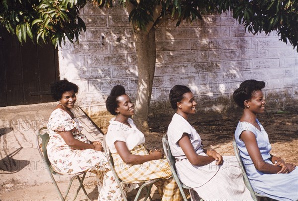Hairstyles of four Ghanaian women. Four female Assistant Education Officers sit in a row in profile, smiling as they model their hairstyles for the camera. Saltpond, Ghana, circa 1958. Saltpond, West (Ghana), Ghana, Western Africa, Africa.