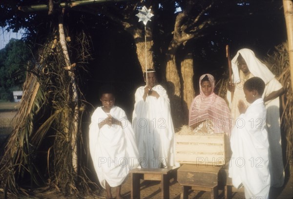 Ghanaian children perform a nativity play. Children in fancy dress perform a nativity play outdoors. Dressed as Mary, Joseph and the angels, they stand with their hands pressed together in prayer beside a straw-filled crate representing the crib of baby Jesus. Ghana, circa 1960. Ghana, Western Africa, Africa.
