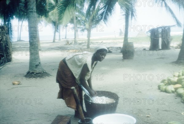 Woman grating a coconut. A woman grates fresh coconut into a basket on the beach at Saltpond. Saltpond, Ghana, 4 July 1960. Saltpond, West (Ghana), Ghana, Western Africa, Africa.