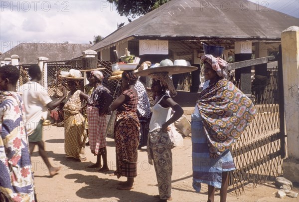 Traders watch an independence procession. Female market traders balance trays of food on their heads as they watch a procession to commemorate the third anniversary of Ghanaian independence. Saltpond, Ghana, circa 1960. Saltpond, West (Ghana), Ghana, Western Africa, Africa.