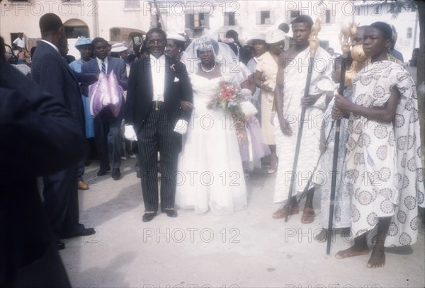 Christian wedding in Ghana. A Ghanaian bride arrives for her wedding accompanied by her father who wears a formal suit. Two men in traditional Ghanaian dress stand nearby holding ceremonial staffs. Ghana, 23 April 1960. Ghana, Western Africa, Africa.