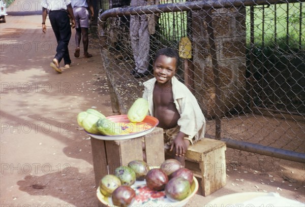 Young fruit seller. A boy sits at a street stall selling fruit to passers-by. Sierra Leone, circa 1960. Sierra Leone, Western Africa, Africa.