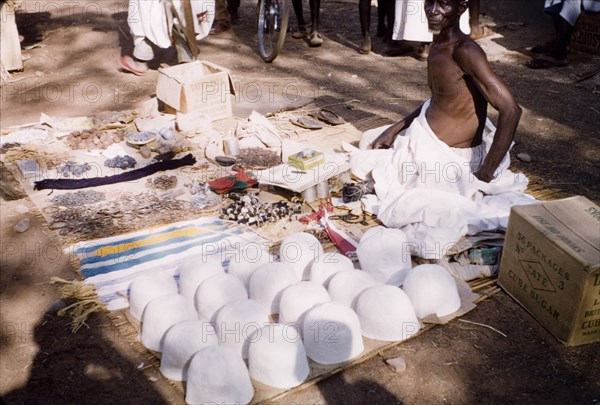 Hat seller at Tamale market. A market trader sits on a woven mat on the ground, selling hats and accessories at an outdoor market. Tamale, Ghana, circa 1961. Tamale, North (Ghana), Ghana, Western Africa, Africa.