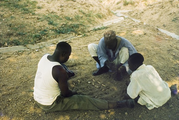 Playing a mancala game, Nigeria. Two men sit on the ground, playing a mancala game in the sand while a young boy looks on. Enugu, Nigeria, circa 1958. Enugu, Anambra, Nigeria, Western Africa, Africa.