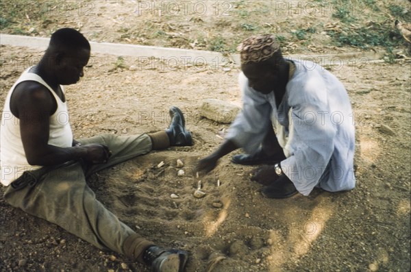 Playing a mancala game, Nigeria. Two men sit on the ground, playing a mancala game in the sand. Enugu, Nigeria, circa 1958. Enugu, Anambra, Nigeria, Western Africa, Africa.